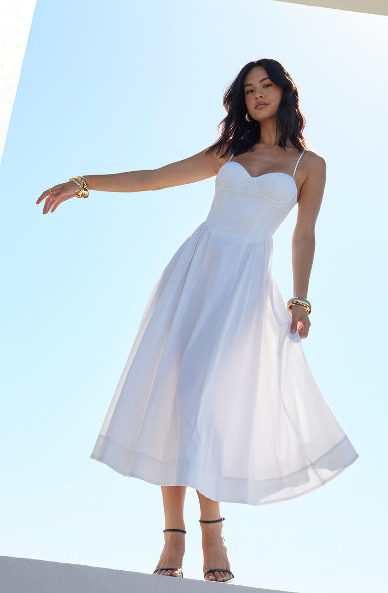 A woman is wearing a white sundress with spaghetti straps, fitted bodice, and a full, flowing skirt that reaches mid-calf. She is accessorized with gold bangles on her left wrist and multiple bracelets on her right wrist, along with gold hoop earrings. She is also wearing black high-heeled sandals with ankle straps.