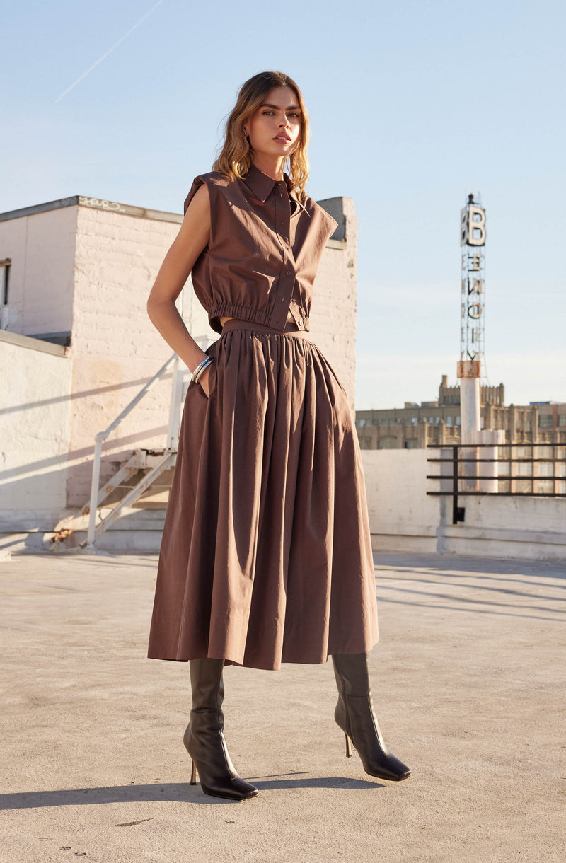 A woman is wearing a sleeveless brown vest with a collar over a dark brown shirt, paired with a high-waisted, full-length brown skirt with gathered fabric. She is also wearing black knee-high heeled boots.