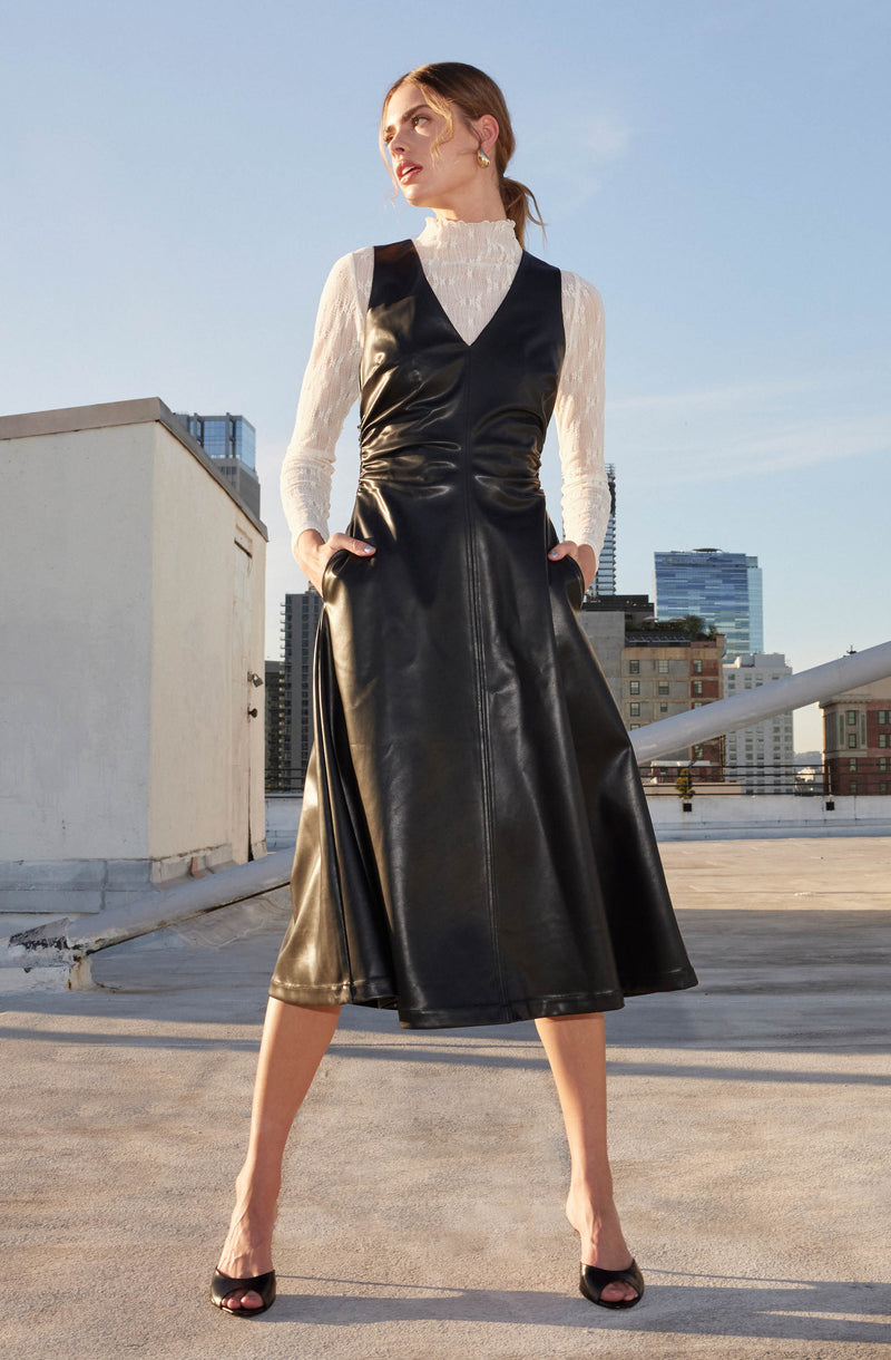 A woman is wearing a white, long-sleeved, high-neck blouse with ruffle detailing at the collar. Over the blouse, she has on a sleeveless, black leather dress with a V-neckline that extends to her ankles. She completes her outfit with black open-toe heels and gold earrings.