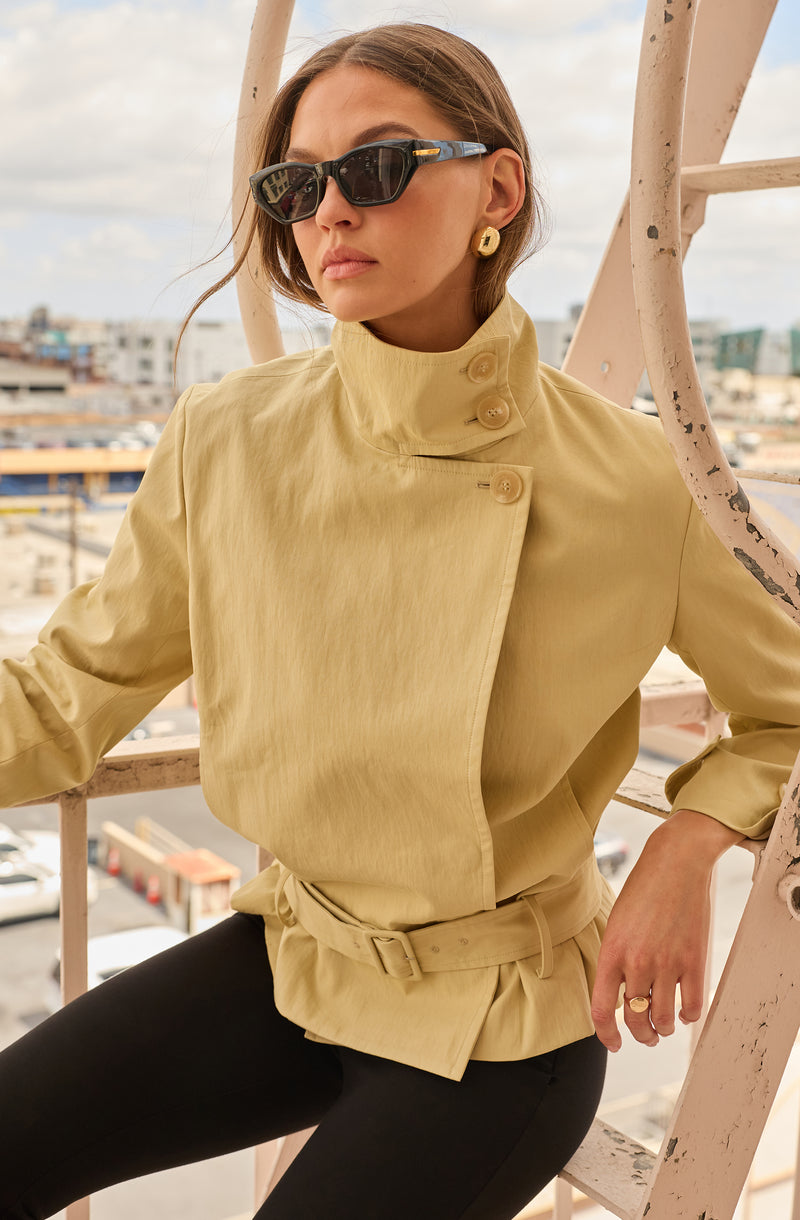 Woman wearing a beige jacket with high collar and large buttons, black pants, and sunglasses.