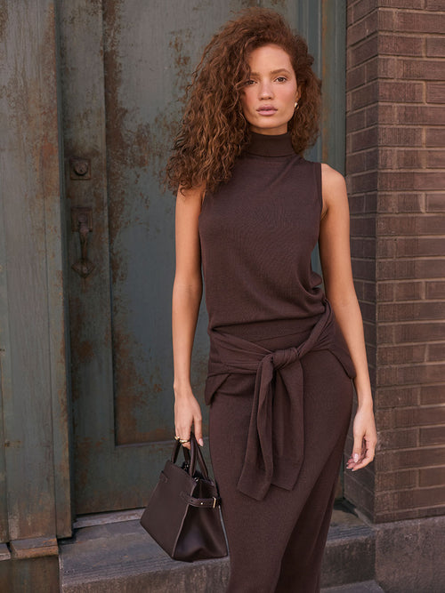 Woman in a sleeveless, high-neck brown outfit with a tied sweater around her waist, carrying a black handbag.