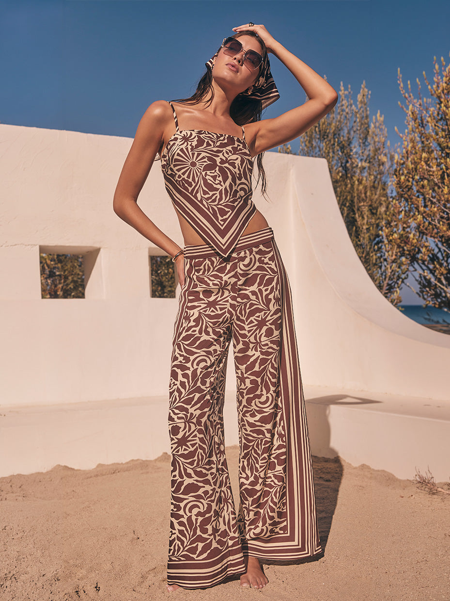 Woman wearing a matching brown and beige patterned sleeveless top and wide-leg pants, sunglasses, and a headscarf, standing outdoors on sand.