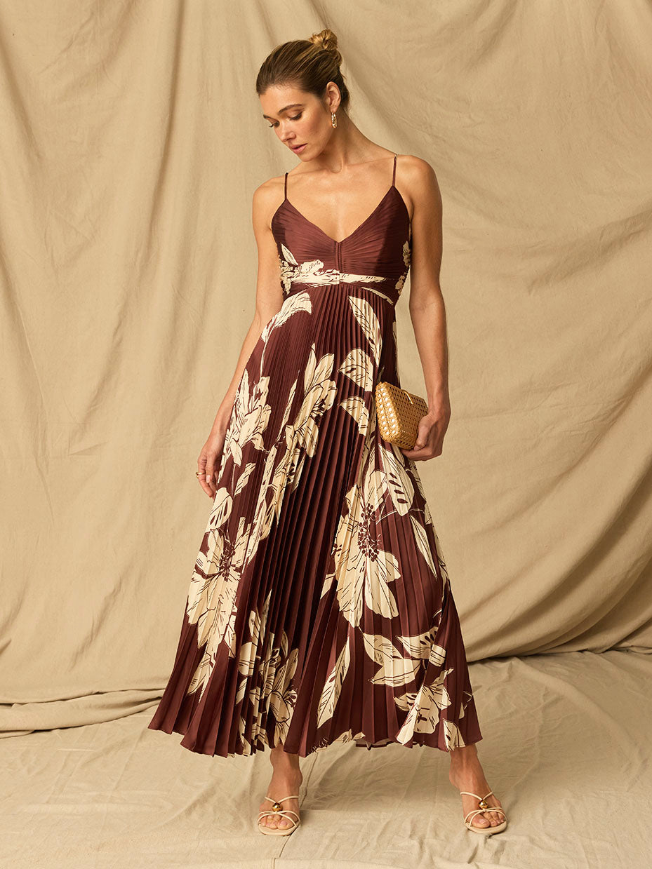 Woman wearing a sleeveless, brown floral pleated dress with spaghetti straps, holding a small beige clutch.