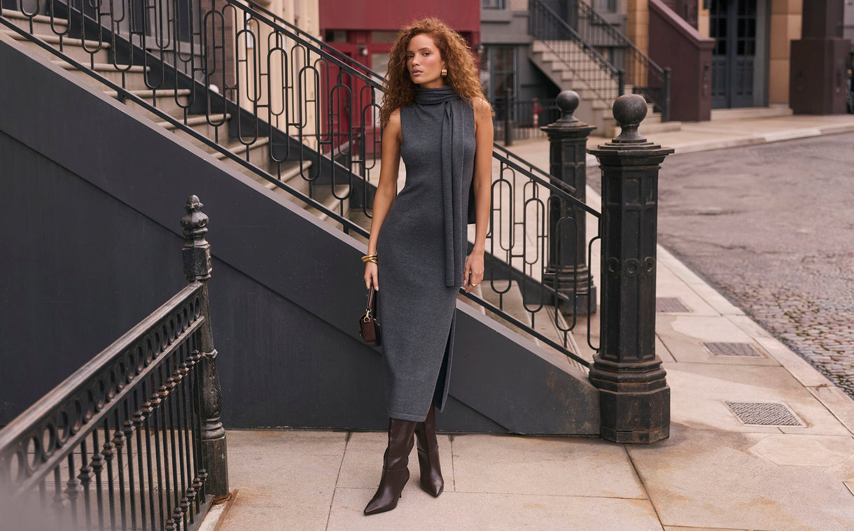 Woman in a sleeveless, dark grey midi dress with side slit and high boots on city sidewalk.