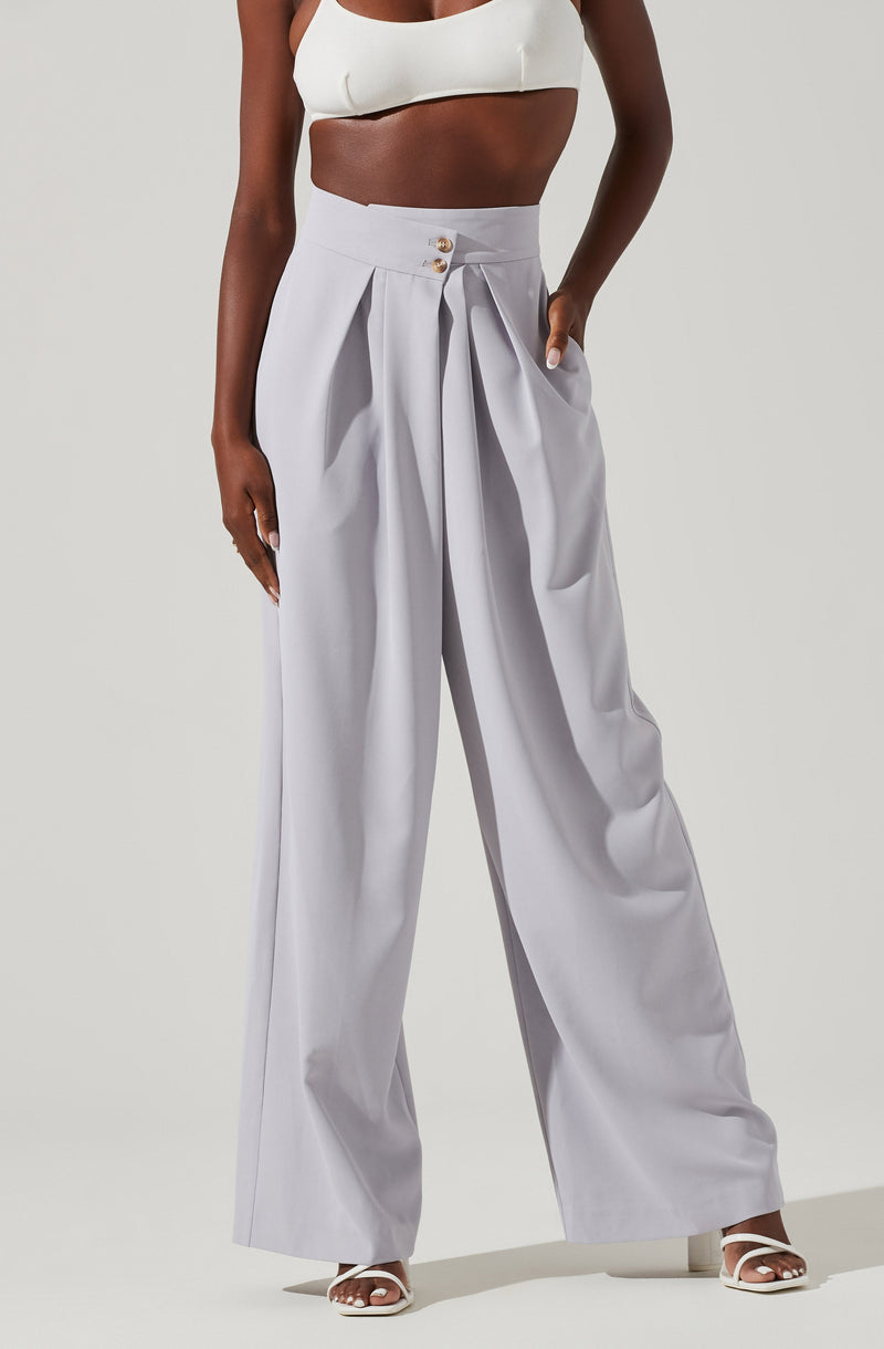 A woman is wearing high-waisted, wide-leg light gray trousers with pleats and a double button closure in the front. She has on a white crop top with thick straps, exposing her midriff. She is also wearing white open-toe heeled sandals.
