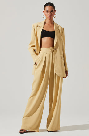 A woman is wearing a light beige tailored pantsuit with wide-leg trousers and a matching blazer. The blazer has a single button closure and a notched lapel collar. She pairs the suit with a black crop top that exposes her midriff. She accessorizes with large gold hoop earrings and black high-heeled sandals.