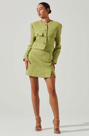 A woman is wearing a matching two-piece outfit consisting of a textured, light green jacket with shoulder pads and four decorative buttons, paired with a matching textured skirt that has a slit on the side. She is also wearing nude-colored, strappy high-heeled sandals.