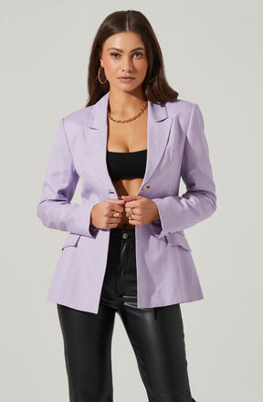 A woman is dressed in a light purple blazer with a notched lapel collar and long sleeves. Underneath, she wears a black crop top, exposing her midriff. She pairs the outfit with high-waisted black leather pants. She accessorizes with large gold hoop earrings, a chunky gold chain necklace, and multiple gold rings.
