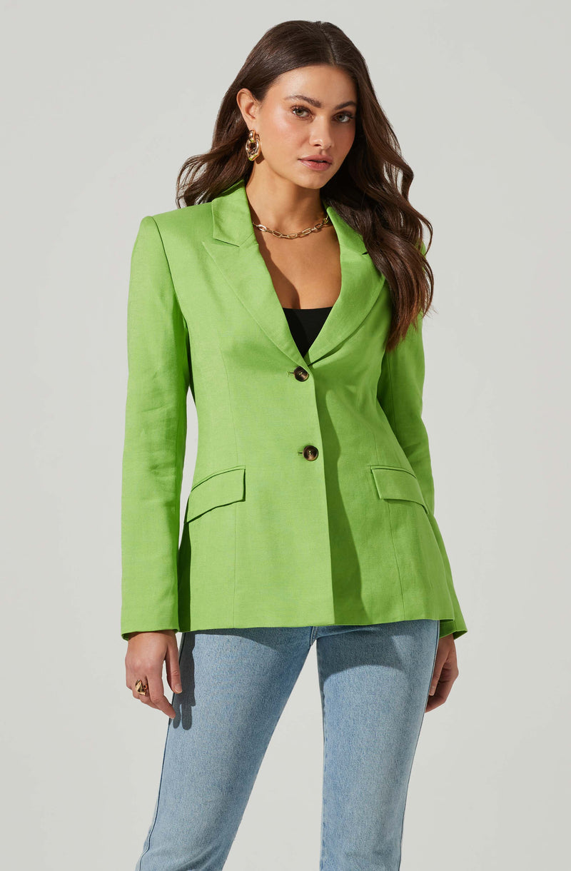A woman is wearing a vibrant lime green blazer with a tailored fit, featuring a notched lapel collar, two front pockets with flaps, and two buttons in the front. She pairs it with light blue jeans and accessorizes with gold hoop earrings, a gold chain necklace, and a ring on her right hand.