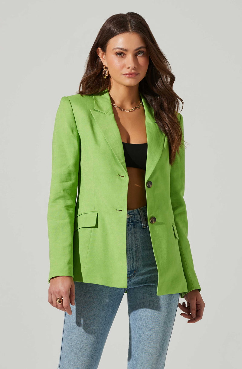 A woman wearing a bright green blazer with a notched lapel, two dark buttons, and flap pockets, paired with a black crop top and light blue jeans. She accessorizes with gold hoop earrings, a gold chain necklace, and a gold ring.