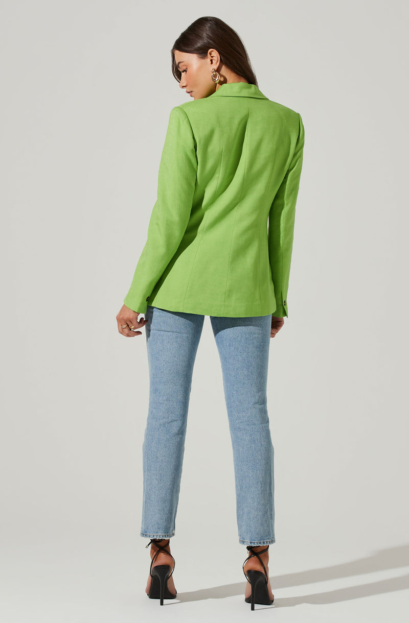 A woman is wearing a bright green blazer with a tailored fit, structured shoulders, and long sleeves. The blazer has a classic lapel collar and buttoned cuffs. She pairs it with light blue, straight-leg jeans and black high-heeled shoes with ankle straps. She accessorizes with gold hoop earrings.