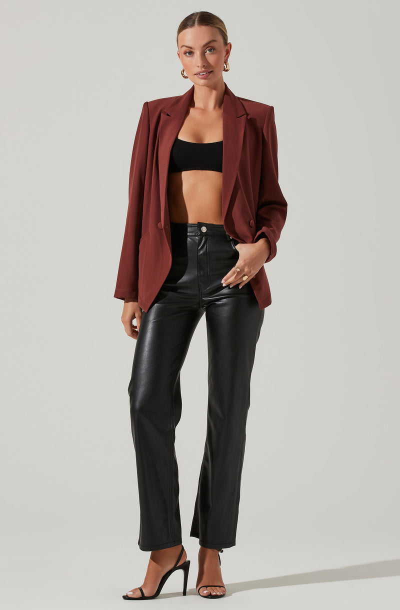A woman is wearing a burgundy blazer with a relaxed fit and slightly padded shoulders over a black crop top. She has on high-waisted black leather pants and black high-heeled sandals.