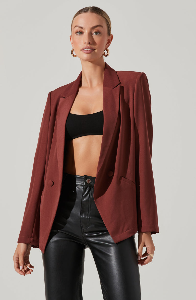 A woman wearing a fitted black crop top, a structured oversized burgundy blazer, and high-waisted black leather pants. She has short hair pulled back, gold hoop earrings, and is styled with minimal makeup.
