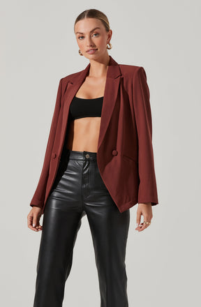 A woman is wearing a rust-colored blazer with structured shoulders and a lapel collar. Underneath, she has on a black crop top that exposes her midriff. She pairs this with high-waisted black leather pants. She accessorizes with gold hoop earrings and rings on her fingers.