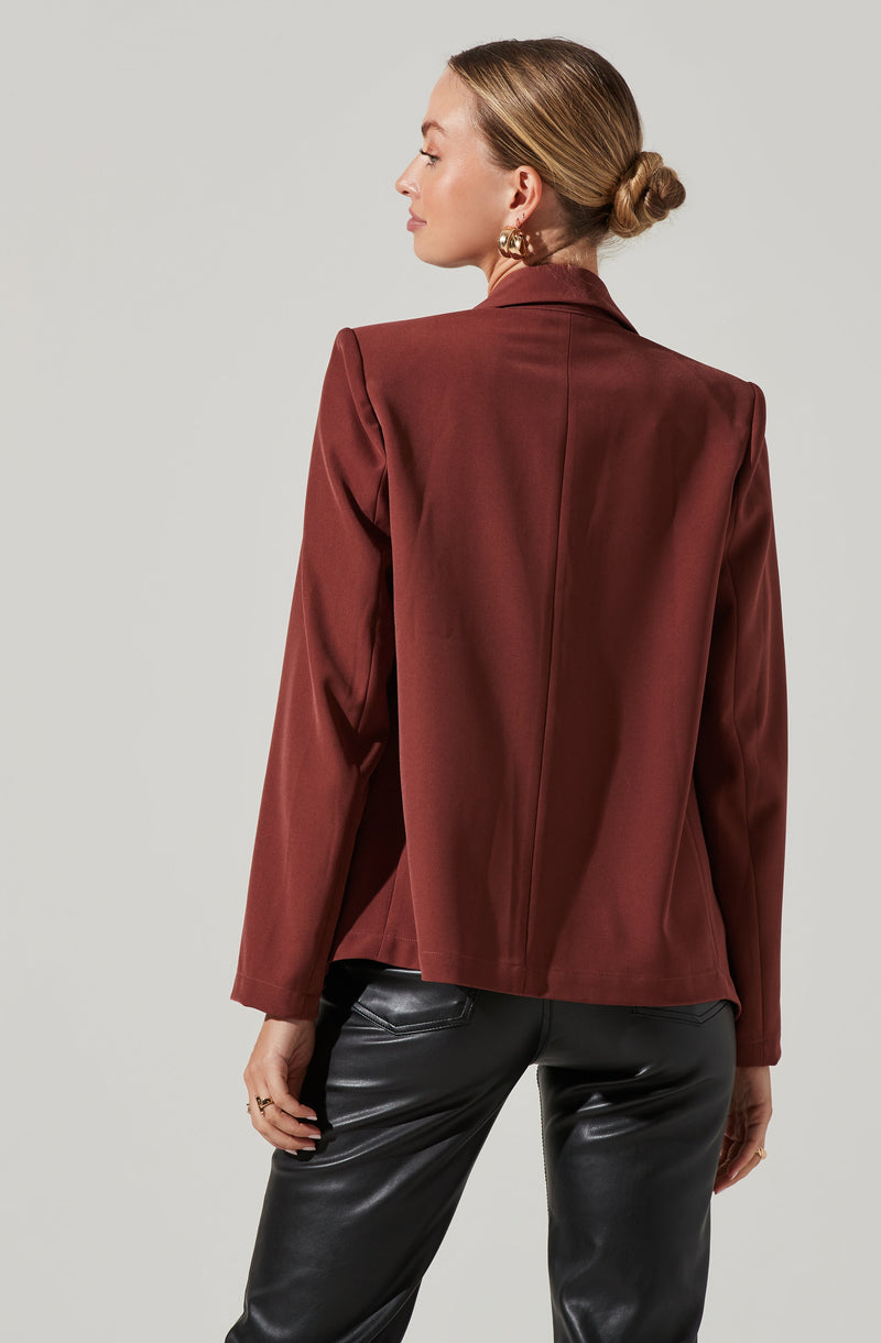 A woman is wearing a burgundy blazer with structured shoulders and a notched collar, paired with black leather pants. She has her hair styled in a low bun and is accessorized with large gold hoop earrings and rings on her fingers.
